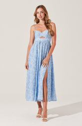 A woman is wearing a light blue, sleeveless dress with thin spaghetti straps. The dress features a fitted bodice with a small cutout below the bust and a high waistline. The skirt is long and flowing, with a high slit on one side revealing her leg. She is also wearing open-toe high-heeled shoes.